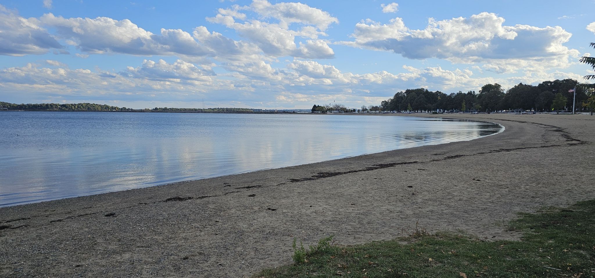 A wide, curving sandy beach meets calm water under a partly cloudy sky. - Boston HarborWalk