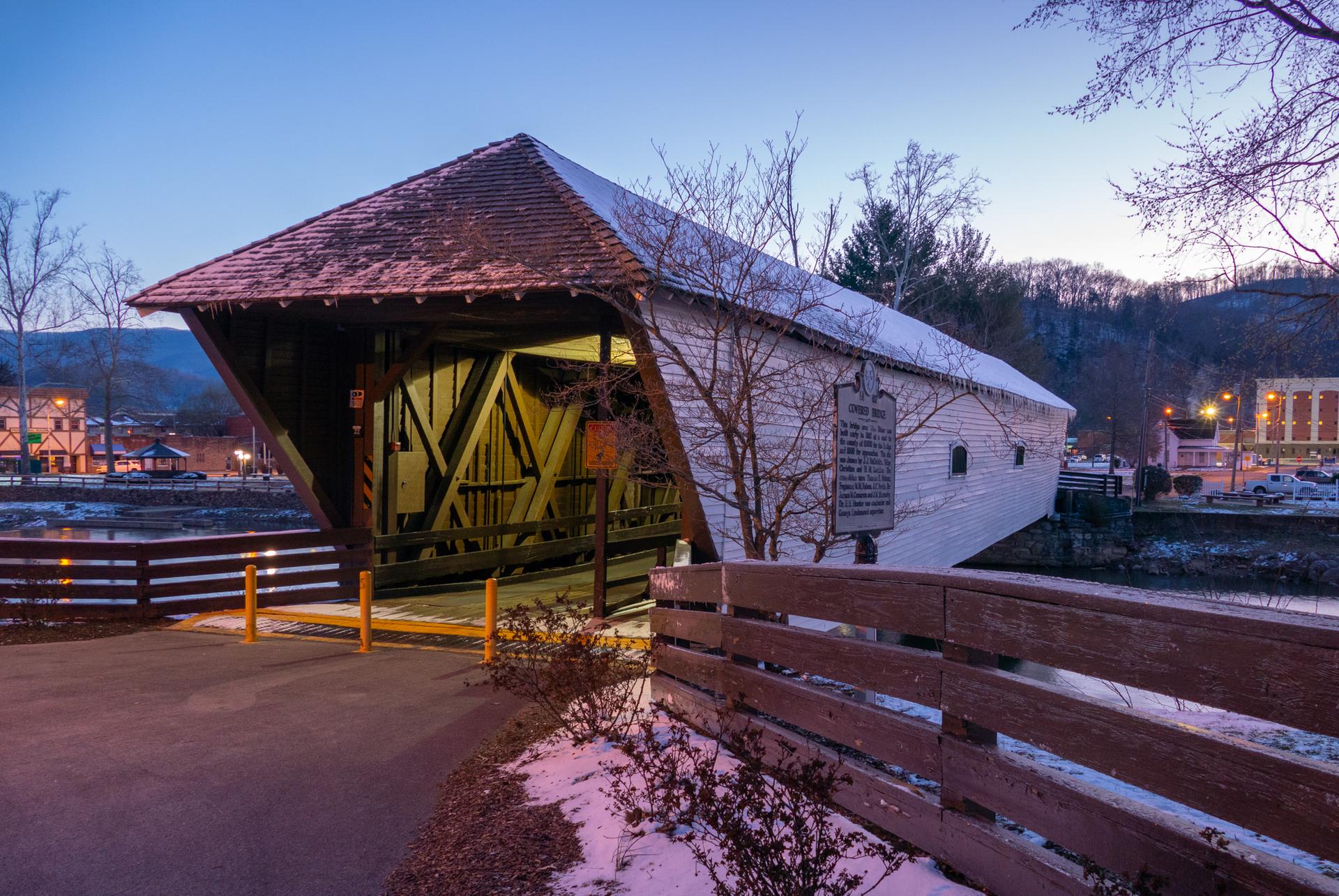 Covered wooden bridge over water at dusk, with snow and distant buildings.