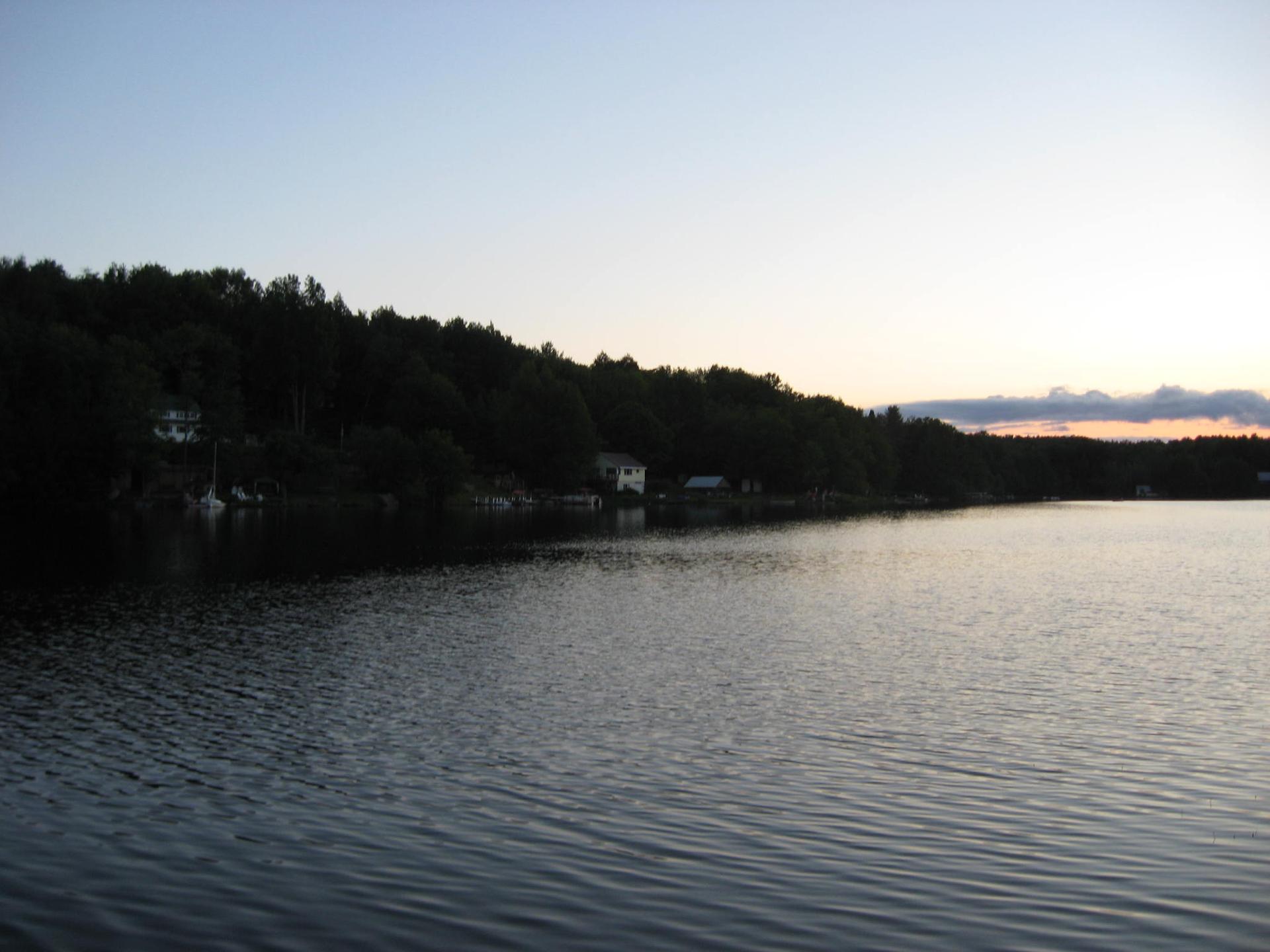 A calm lake at dusk with a forested shoreline and distant houses.