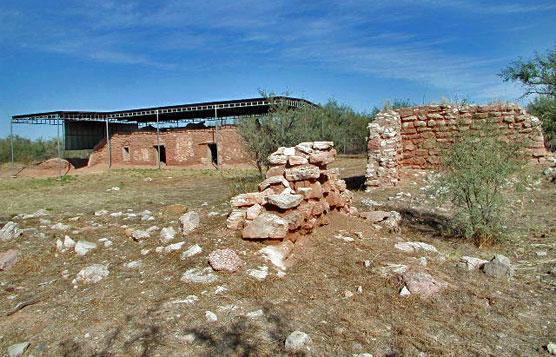Ancient stone and adobe ruins with a modern shelter under a blue sky.