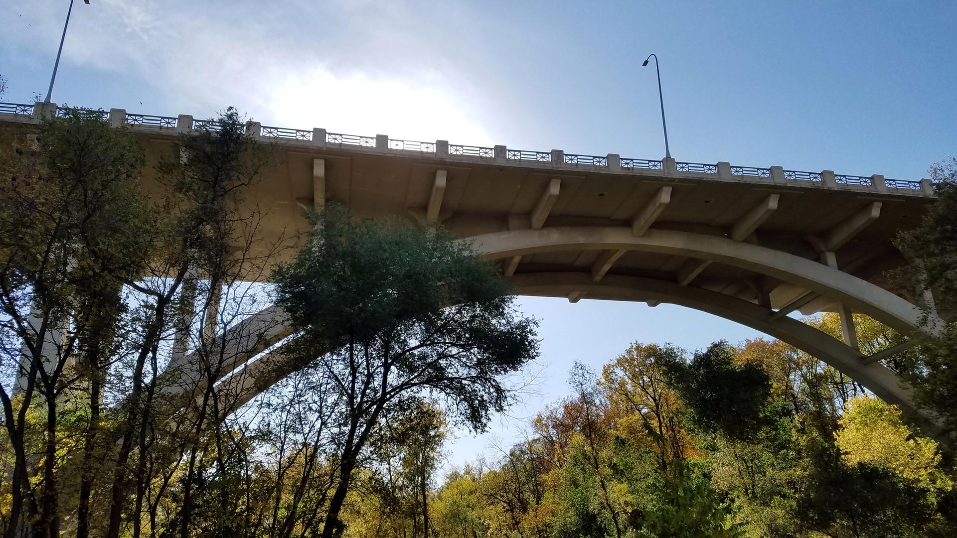 Large concrete arch bridge seen from below, with trees and bright sky.