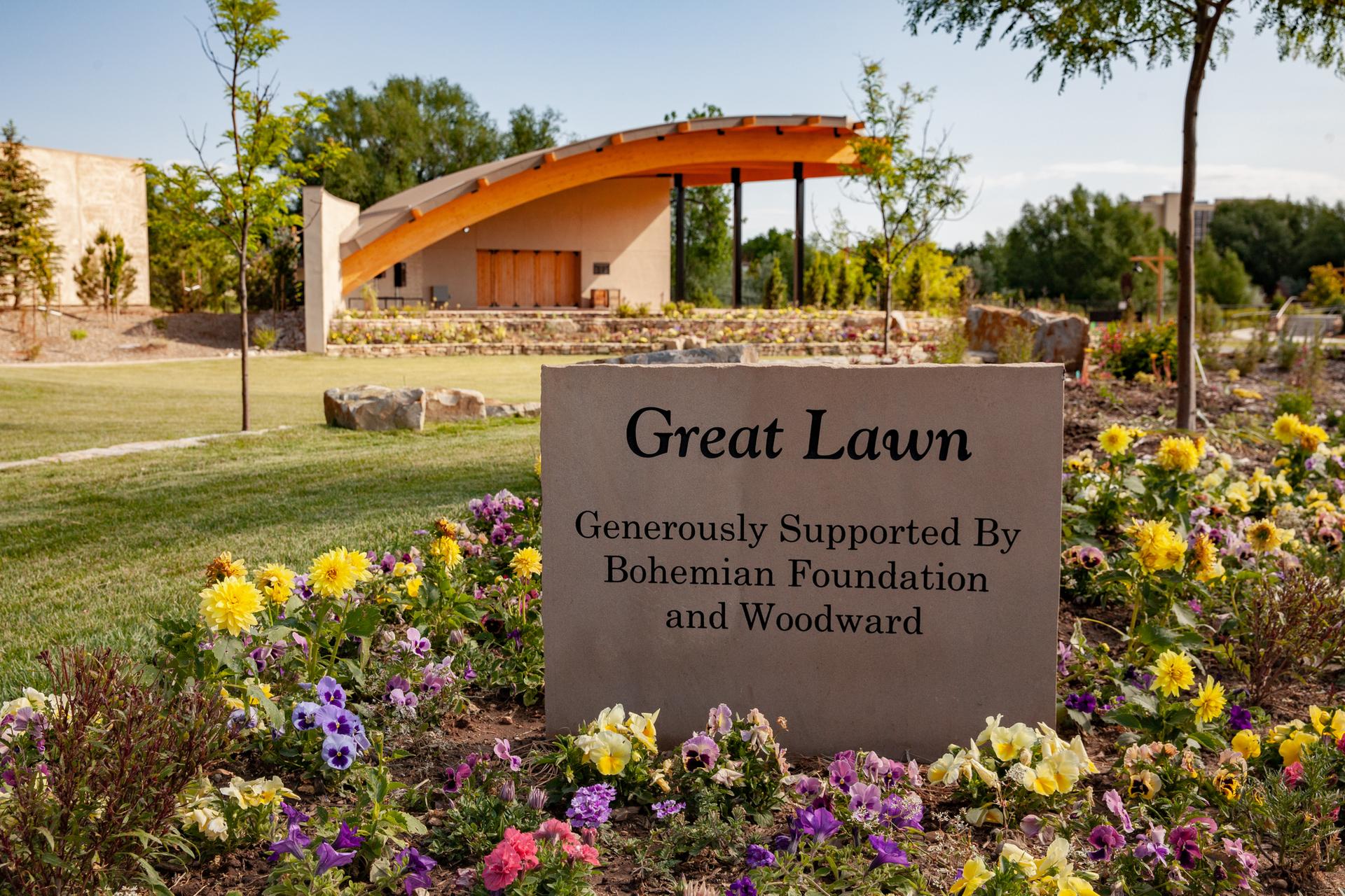 A stone sign surrounded by colorful flowers with a modern building in the background.