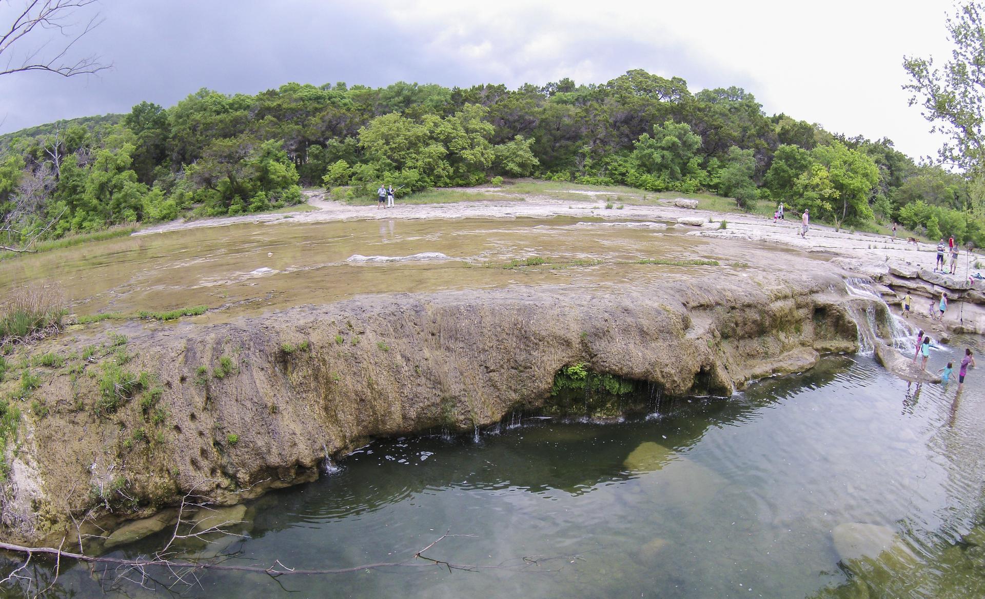 People wading and walking on rocky ledges by a river under cloudy skies.