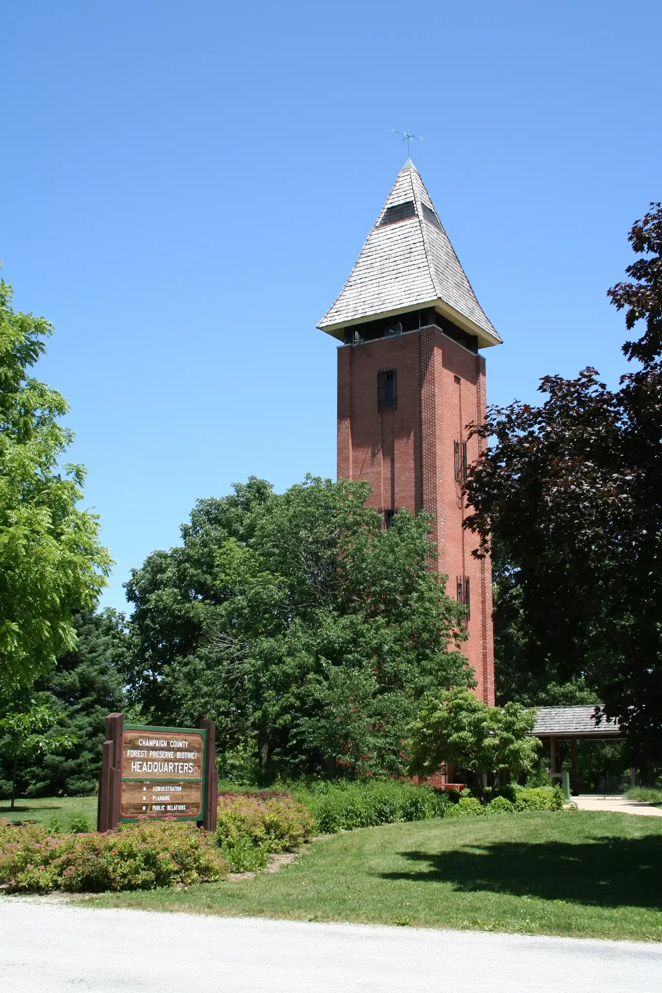 Tall brick tower with a pointed roof, surrounded by green trees on a sunny day.
