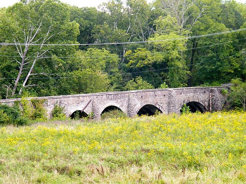 An old stone arch bridge spans a field with yellow wildflowers under bright skies.