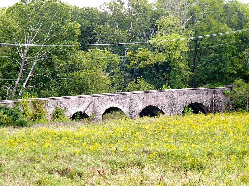 An old stone arch bridge spans a field with yellow wildflowers under bright skies. - Potomac Heritage National Scenic Trail