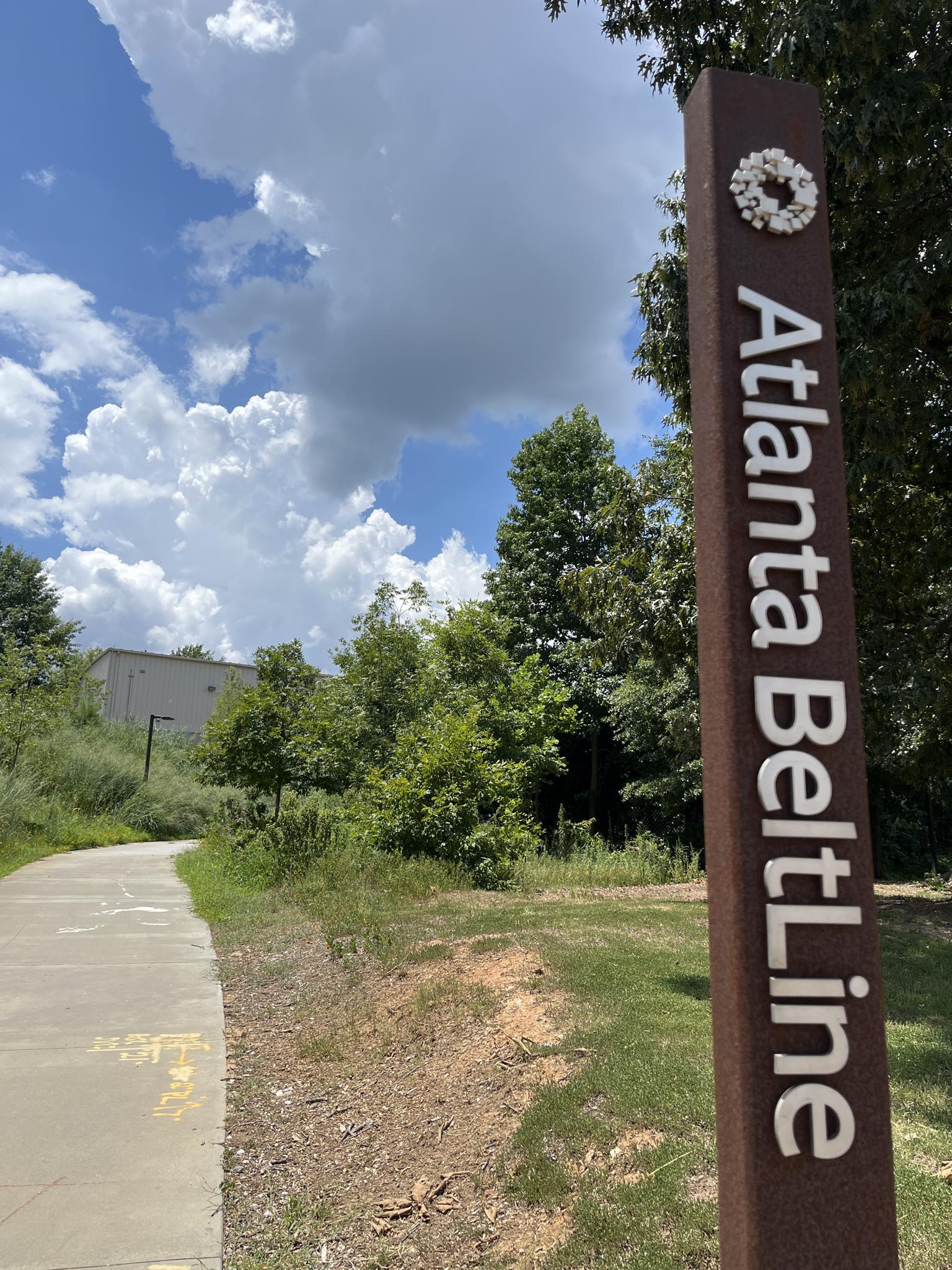 A brown sign stands beside a paved path, surrounded by green trees under a cloudy sky.