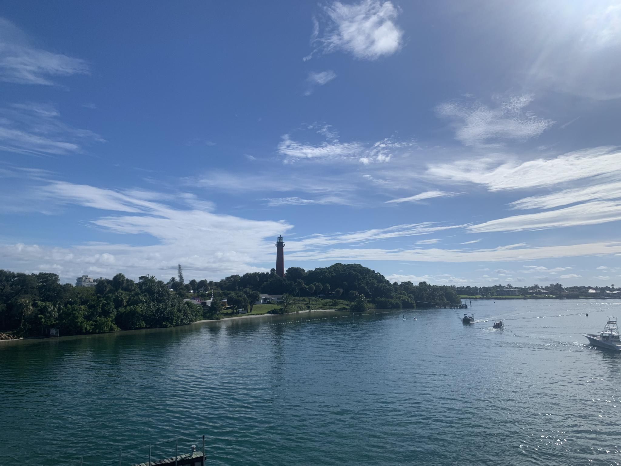 A distant lighthouse overlooks a sunny waterway with boats and green trees. - Jupiter Inlet Lighthouse ONA North Trail