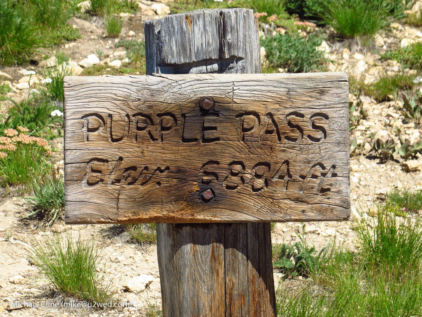 A weathered wooden sign with carved text stands in a sunny, grassy landscape.