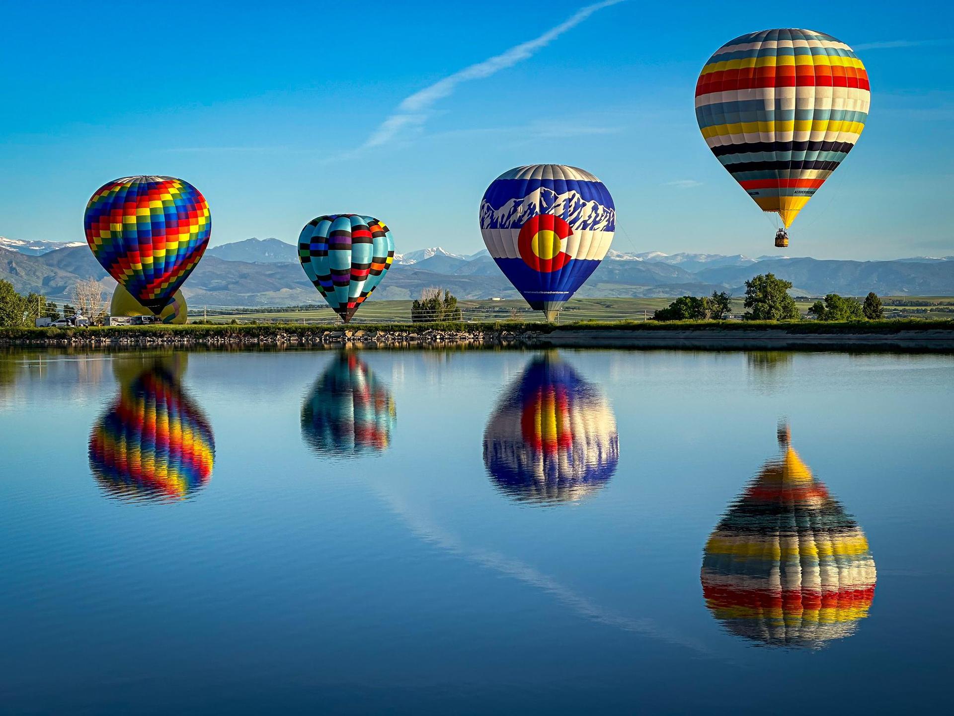 Four colorful hot air balloons reflected in calm water with mountains.