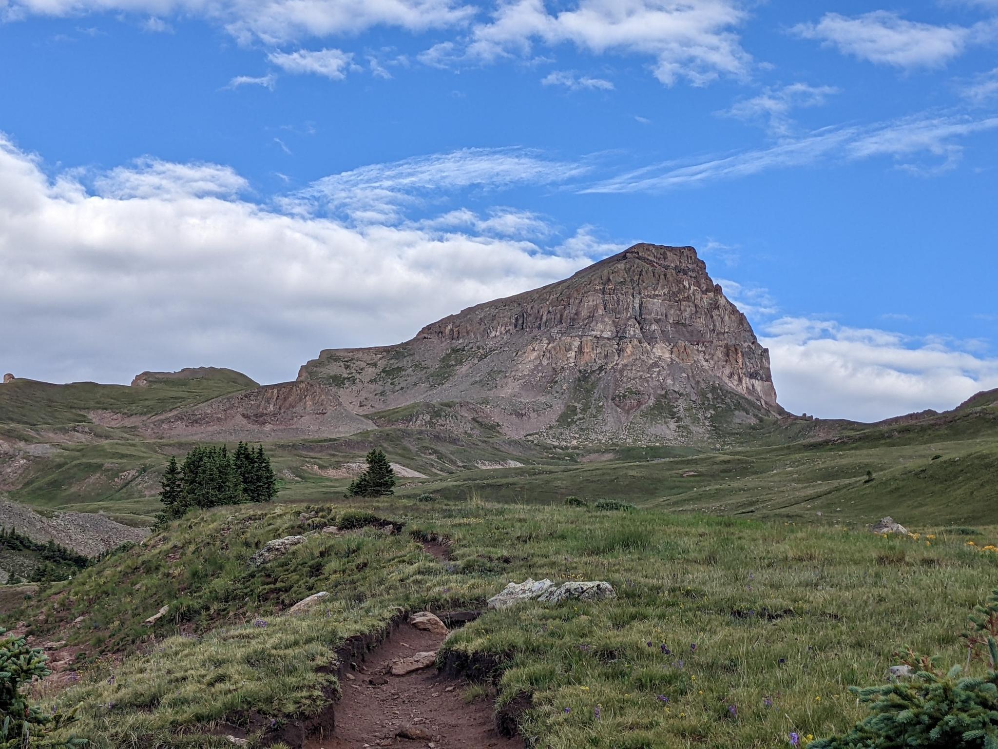 A dirt trail leads through green hills towards a large mountain under blue sky.