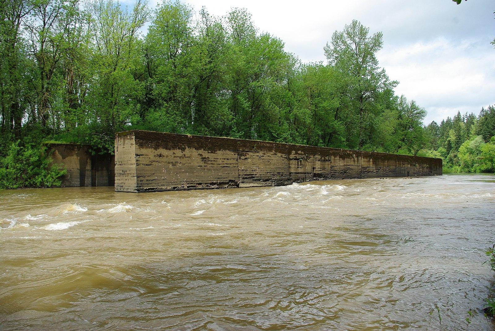 A muddy river flows past a long concrete wall and green trees.