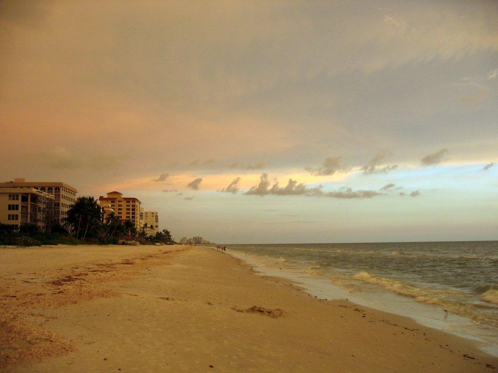 Wide sandy beach with ocean waves and distant buildings under cloudy sky.