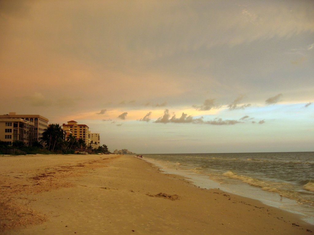 Wide sandy beach with ocean waves and distant buildings under cloudy sky. - Vanderbilt Beach loop from Naples Park
