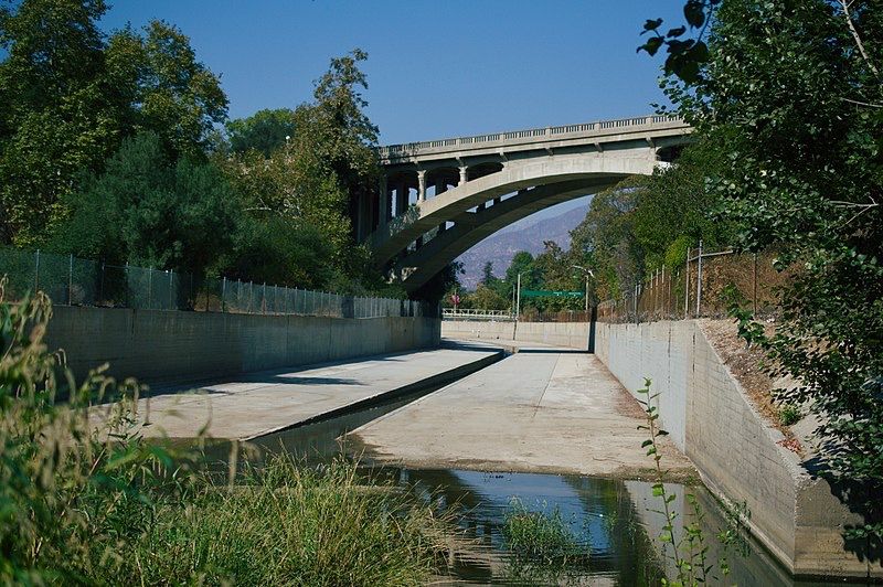 A concrete channel with water flows under a large arched bridge, sunny. - Lower Arroyo Seco Trail