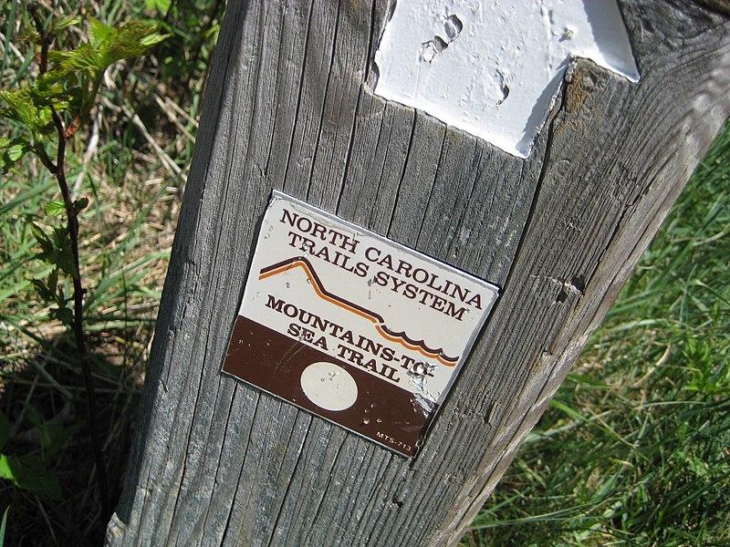 A brown and white trail marker sign on a weathered wooden post.