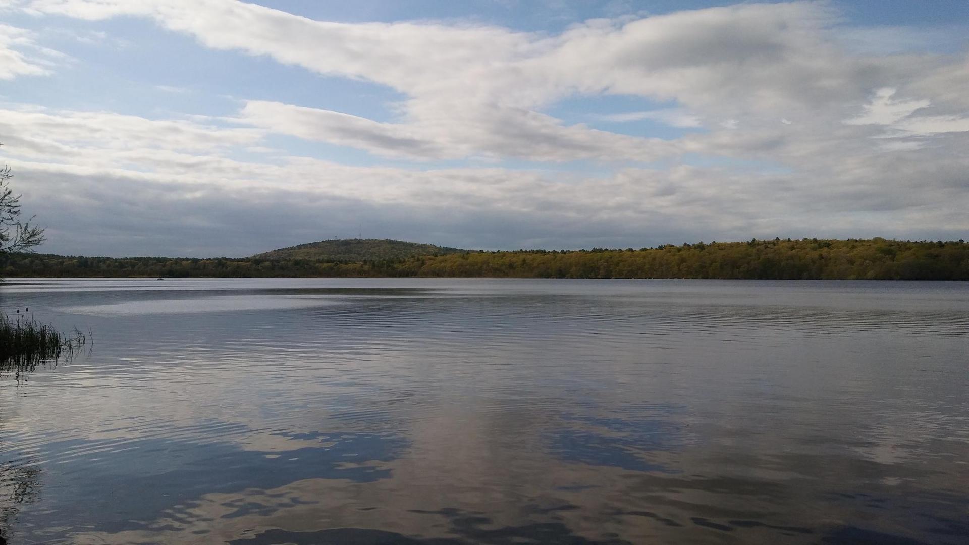 Calm lake reflecting a cloudy sky, with forested hills in the distance.