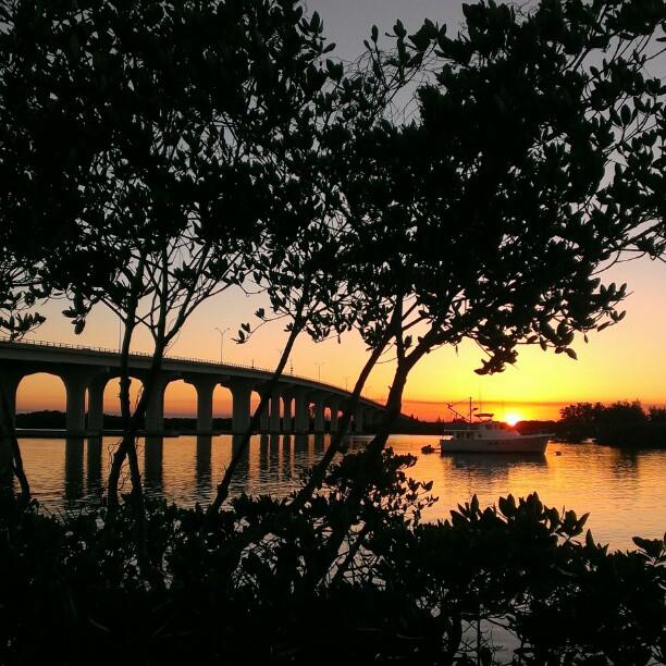 Sunset over water with a bridge, boat, and silhouetted trees.