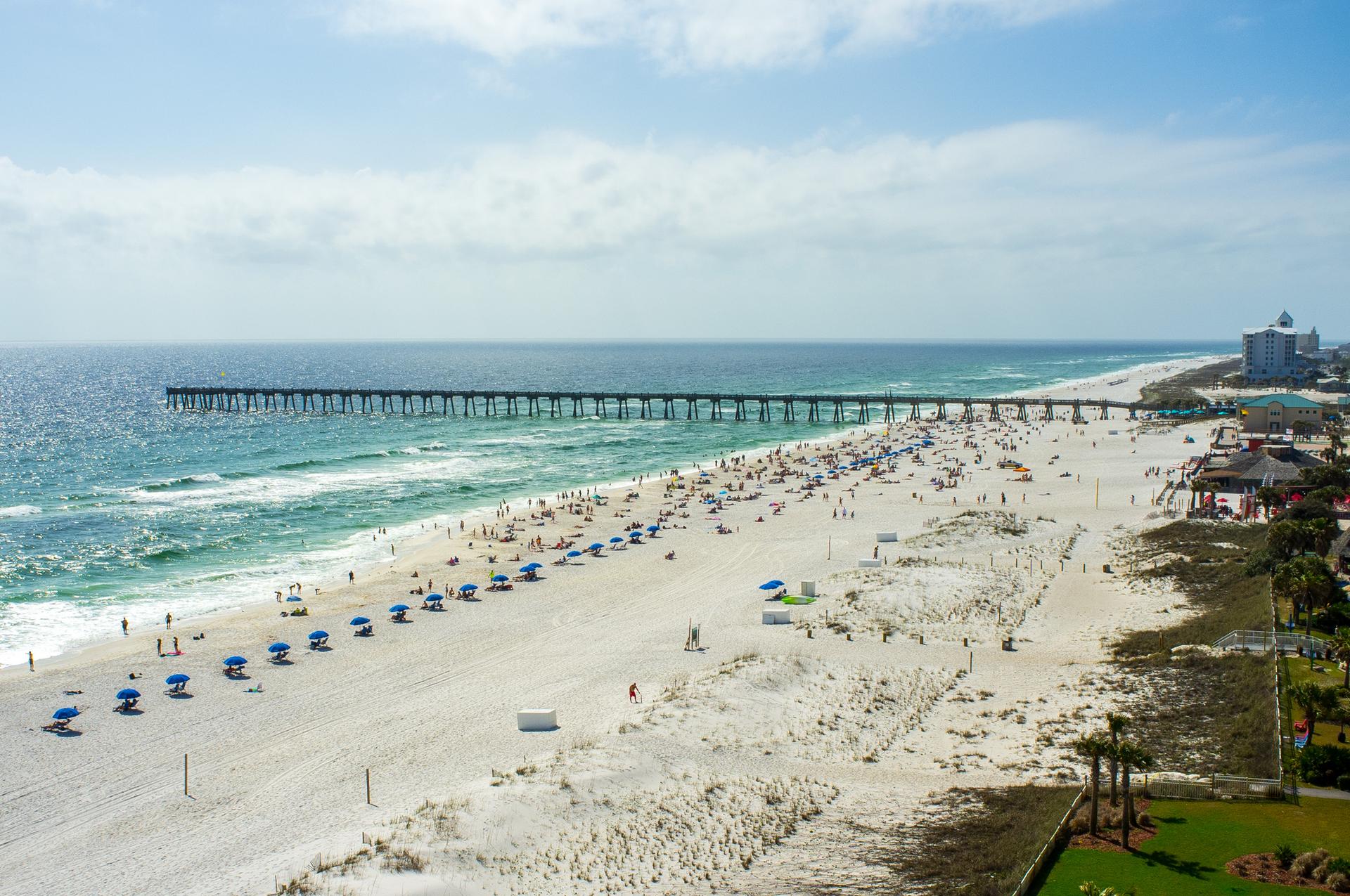 Aerial view of a crowded sunny beach with a long pier and blue umbrellas.