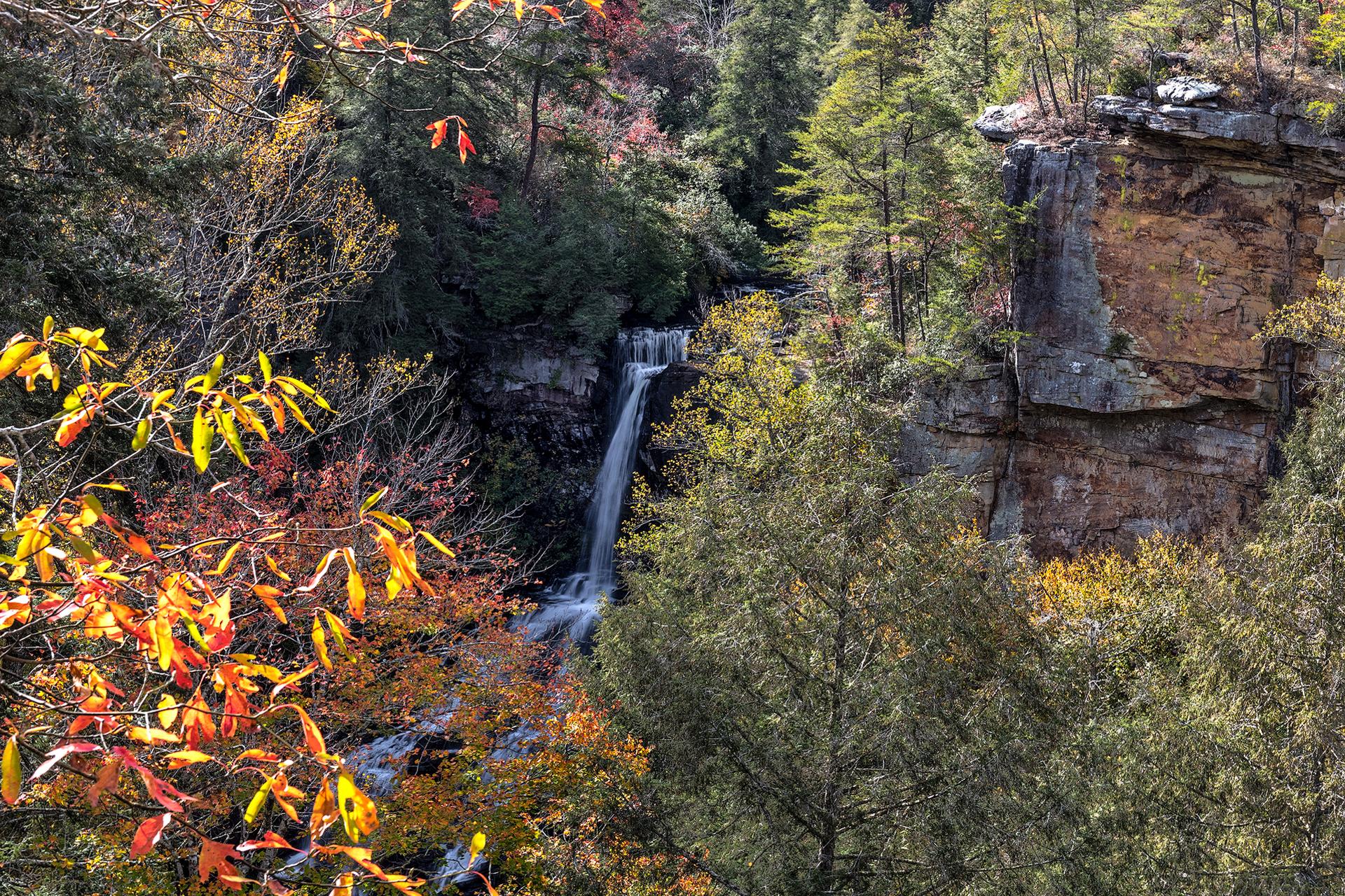 A waterfall cascades down rocky cliffs surrounded by vibrant autumn trees in sunlight.