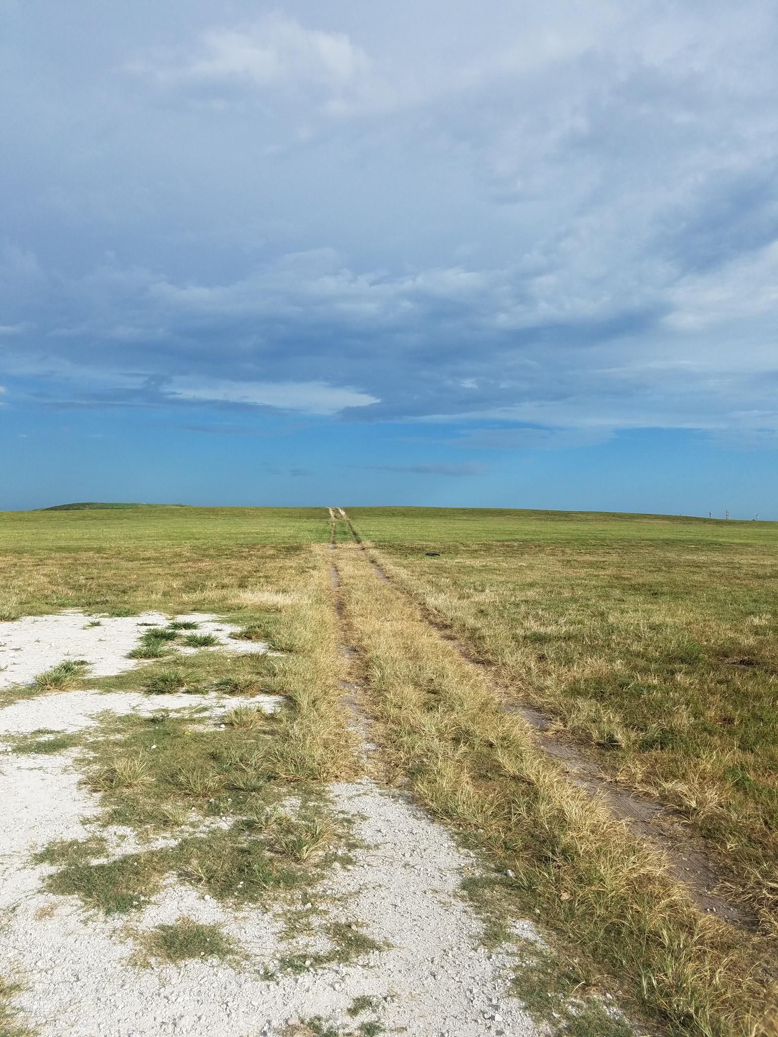 A dirt track extends through a vast, sunny, grassy field under a cloudy sky. - Dyer Perimeter Trail