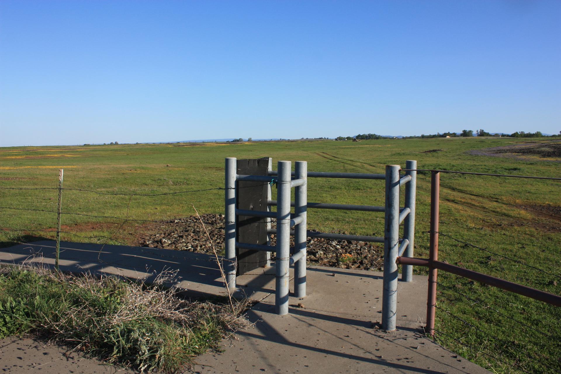 Metal gate in a vast green field under a clear blue sky.