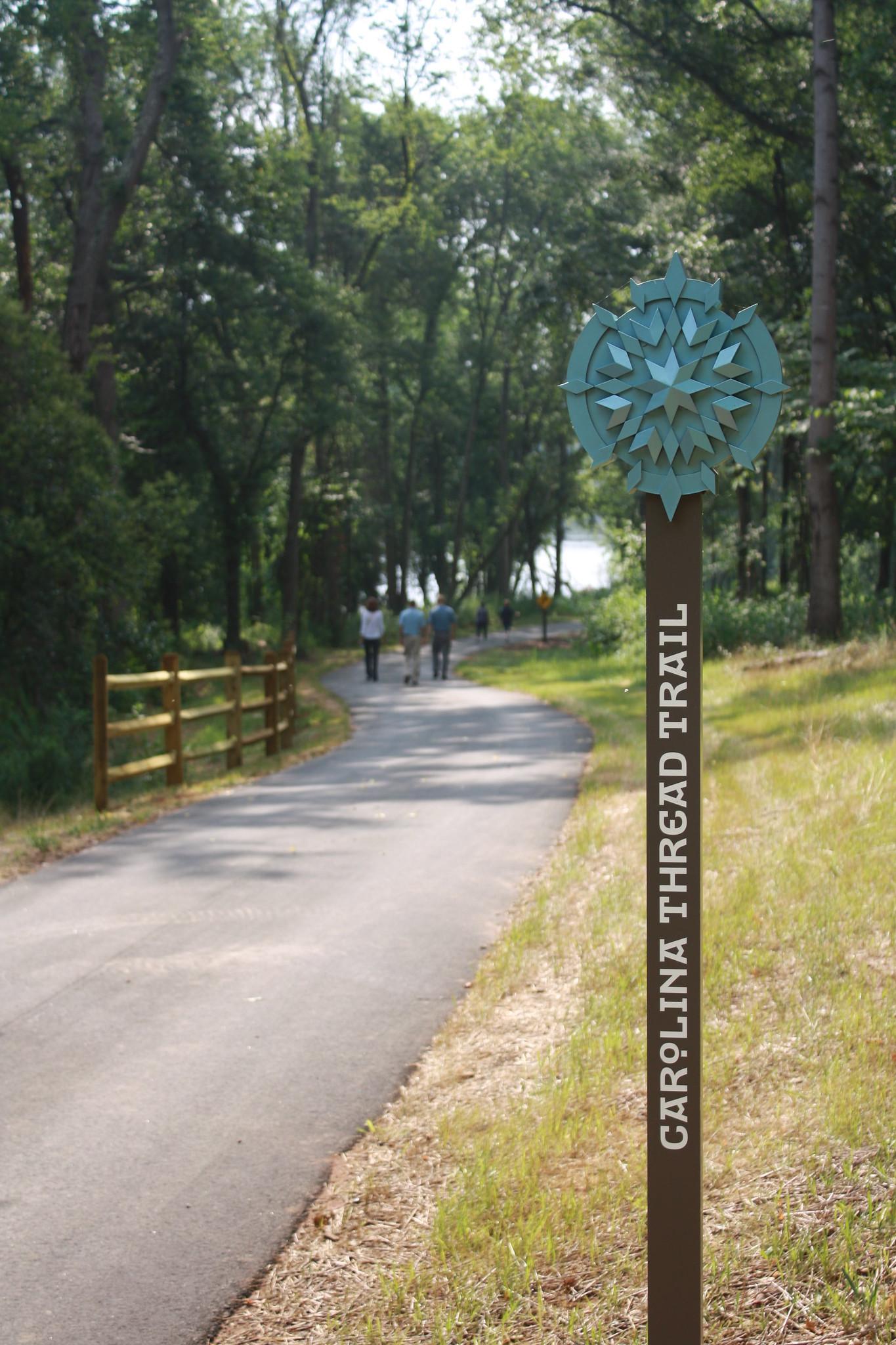 Paved trail through a sunlit forest with a trail marker and people walking.