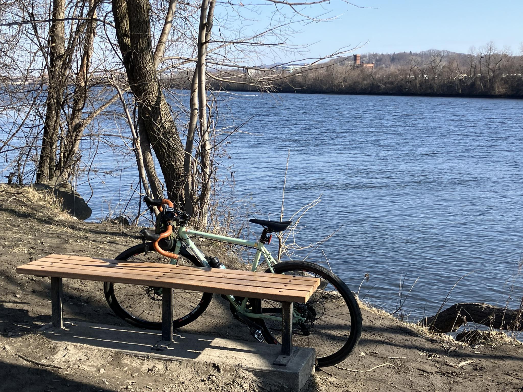 Bicycle parked next to a wooden bench by a wide river on a sunny day.