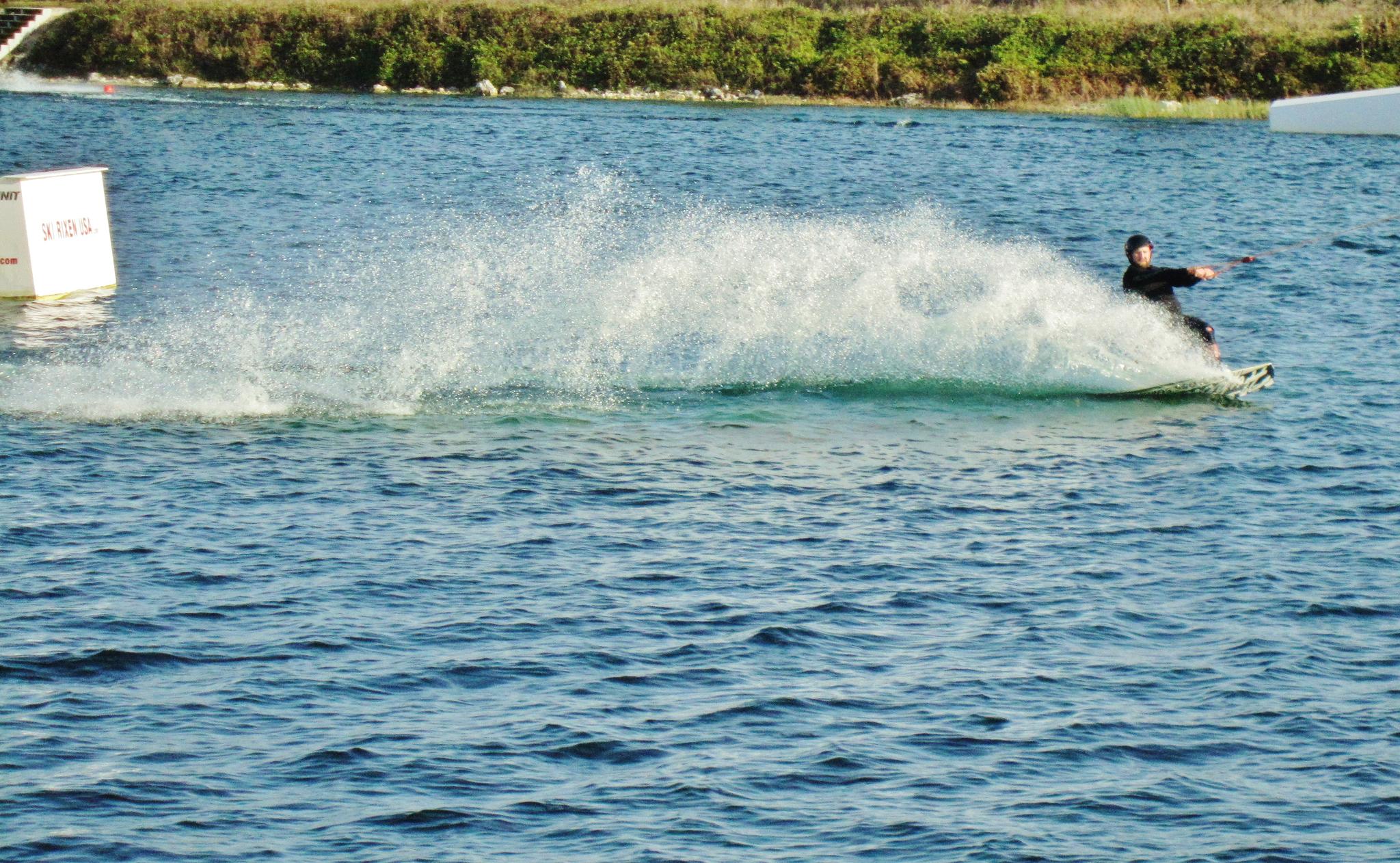 Person waterskiing on blue water, creating a large spray under bright daylight.