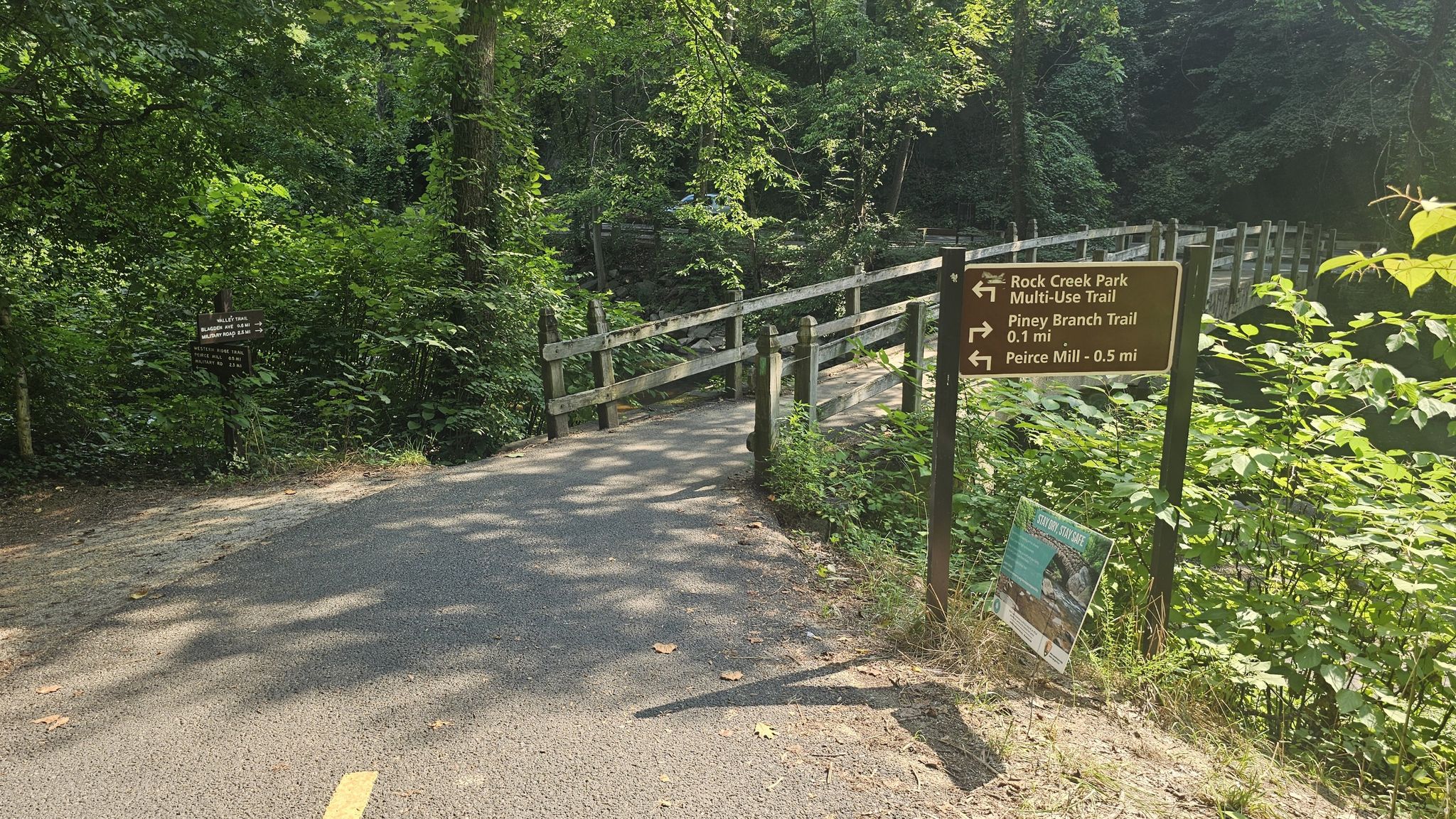 A paved path leads to a wooden bridge and a trail sign in a forest. - Rock Creek Park Loop: Western Ridge and Valley Trails