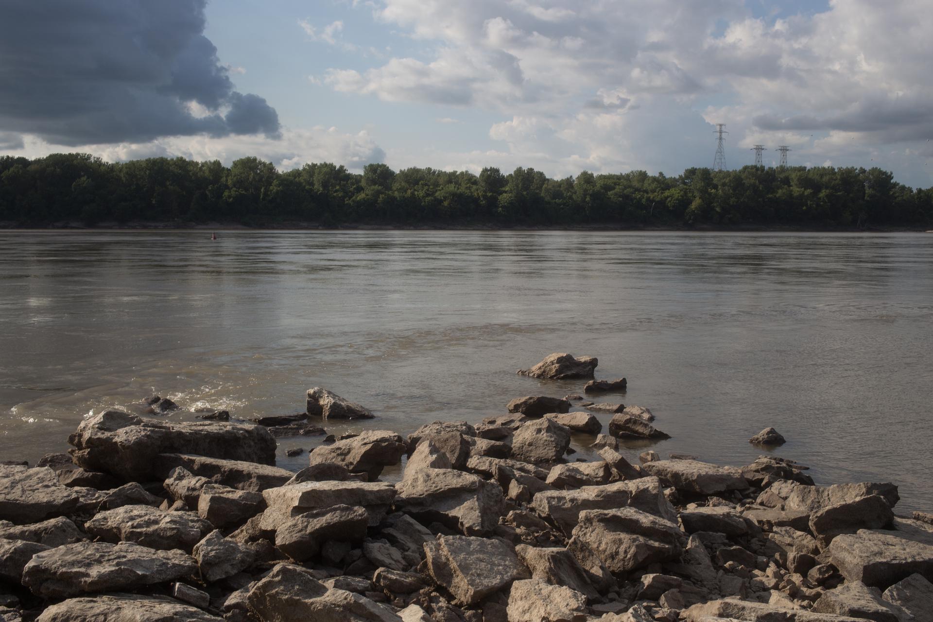 Rocky riverbank with a wide river, distant trees, and cloudy sky.