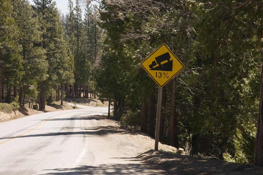 A steep grade warning sign on a paved road through a dense forest.