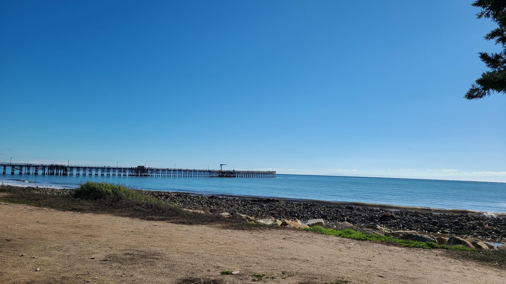 A long pier stretches into the blue ocean from a rocky beach on a clear day. - University of California to Goleta Beach Park run