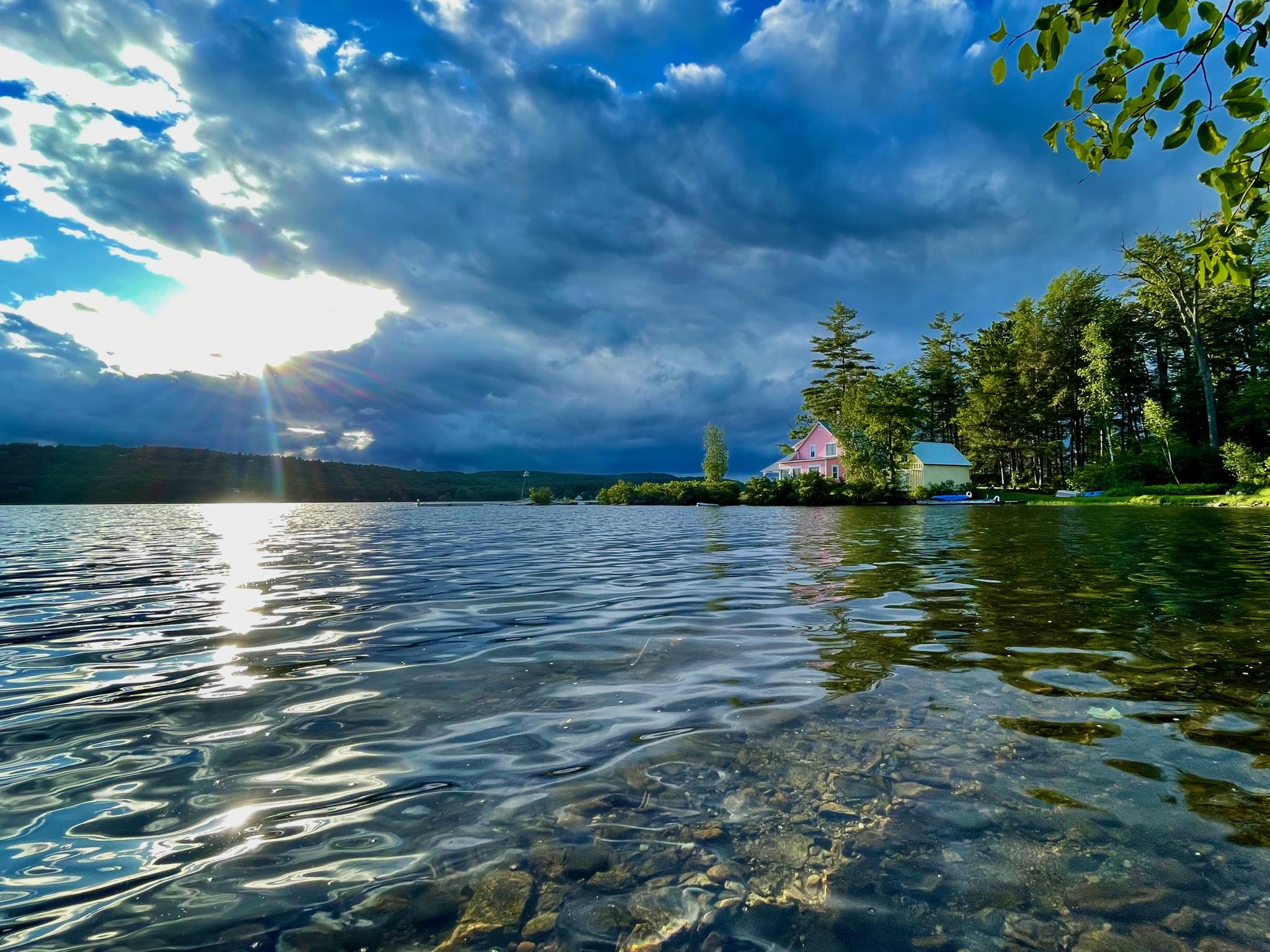 Sunbeams pierce dark clouds over a lake with distant houses and trees.