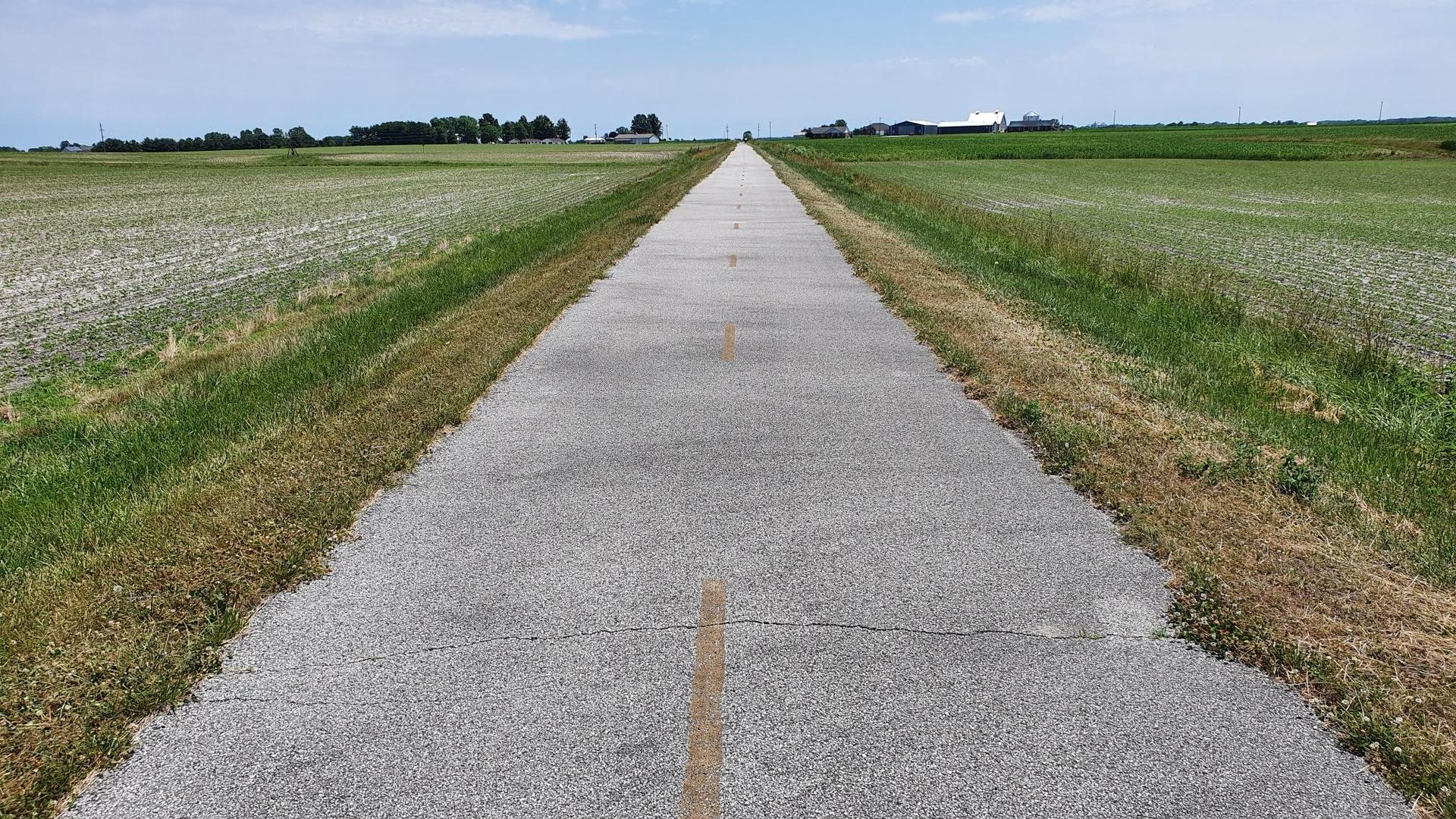 A long, straight paved path extends through flat green fields under a cloudy sky.