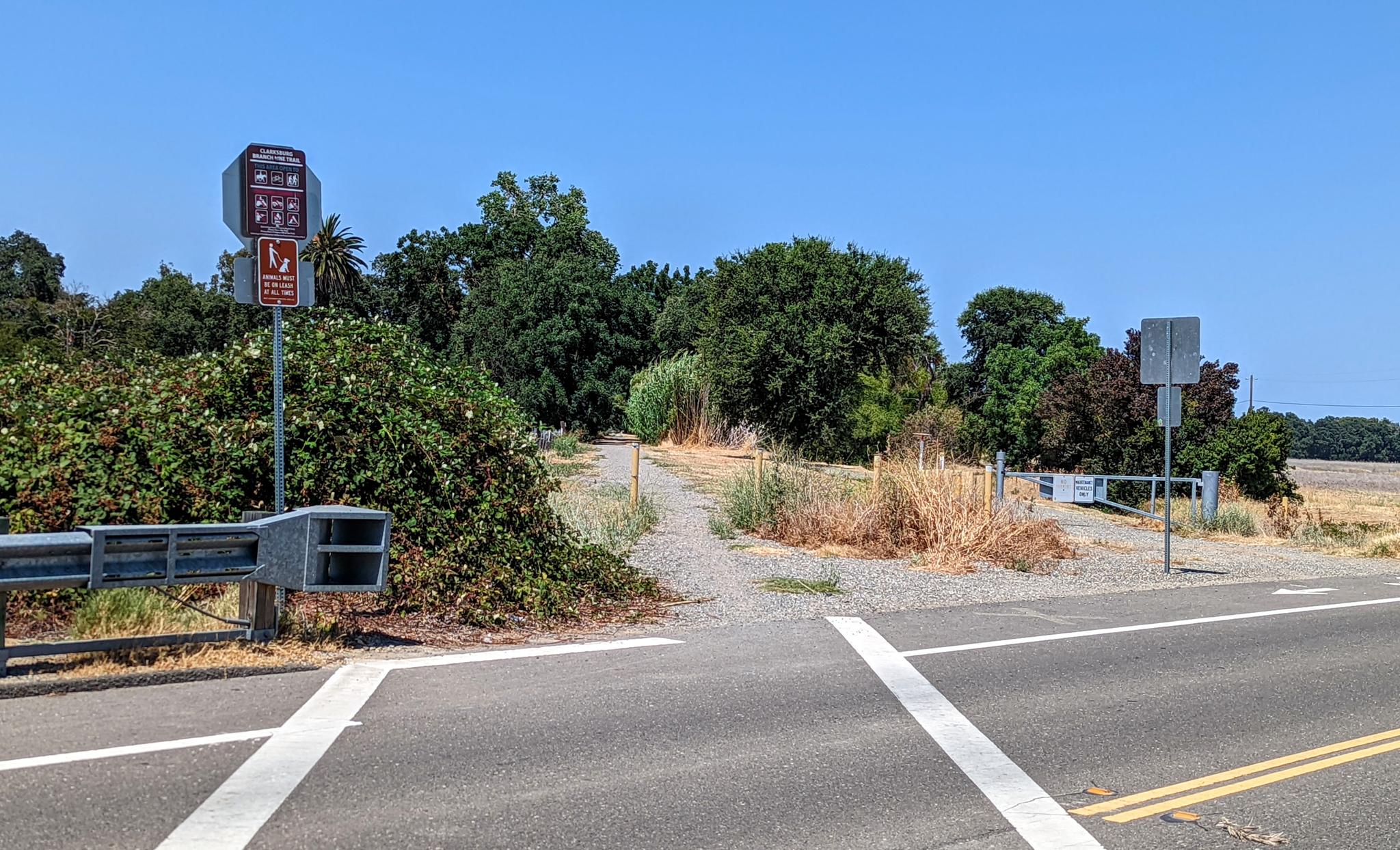 A sunny dirt path entrance next to a paved road with signs.