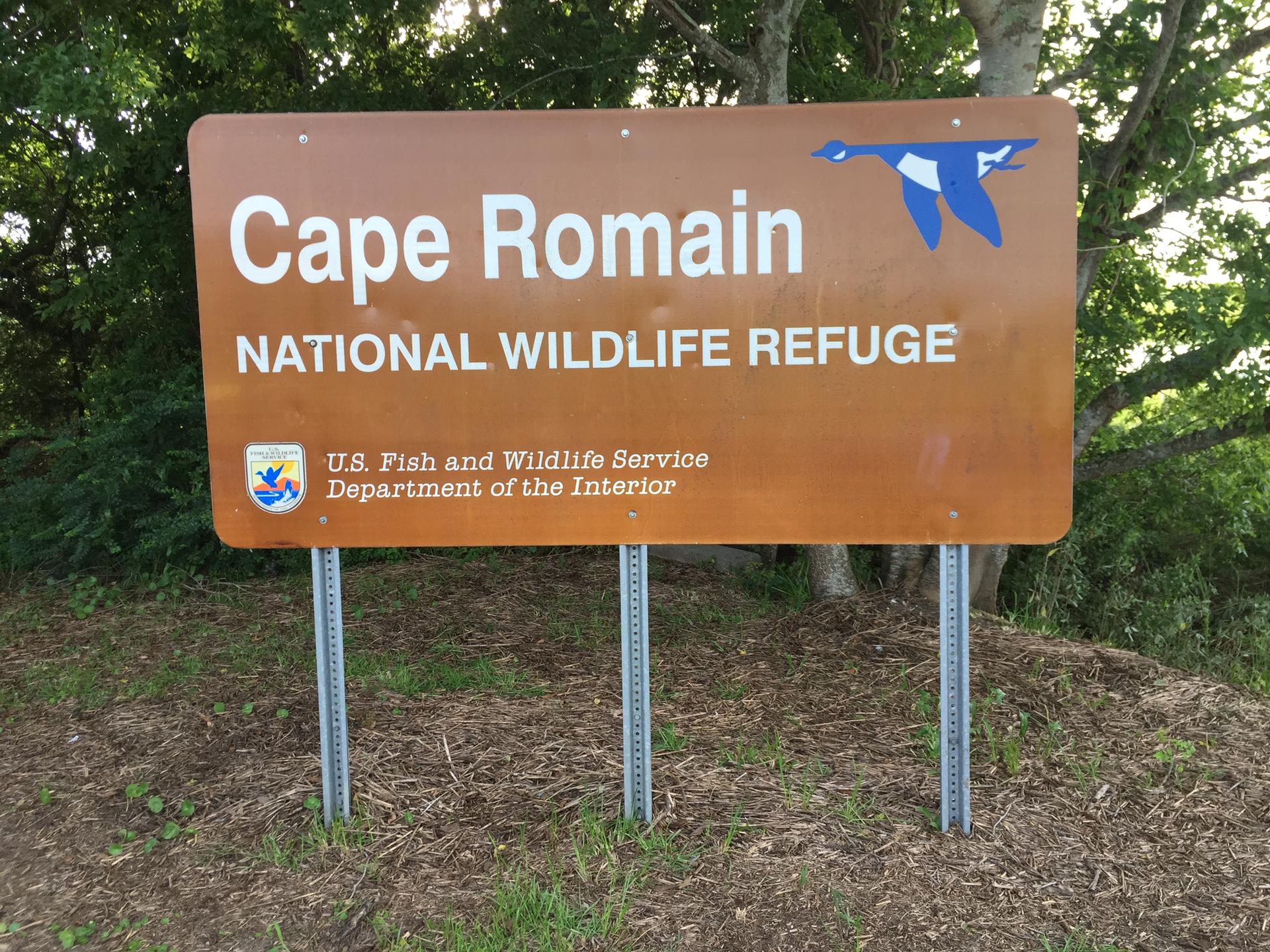 A brown outdoor sign with white text stands in front of green trees.