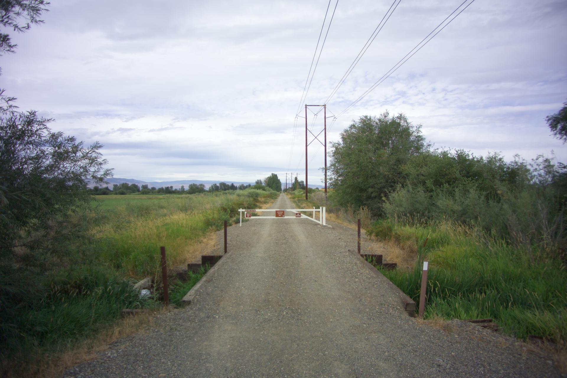 Gravel road blocked by a white gate under power lines, overcast day.