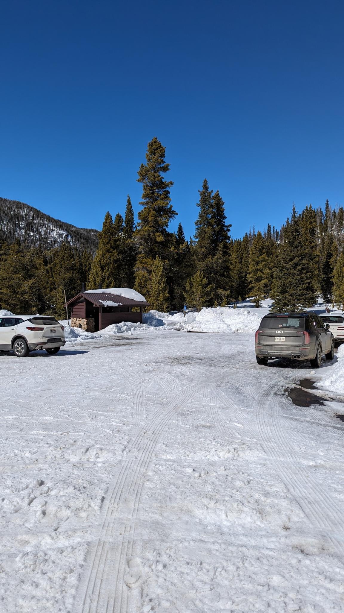Snowy parking lot with cars, a small building, and pine trees under a clear sky.