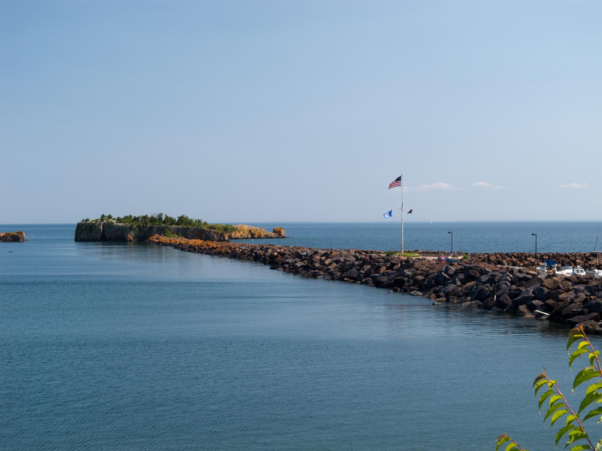 A rocky breakwater with flags extends into calm water under a clear sky.