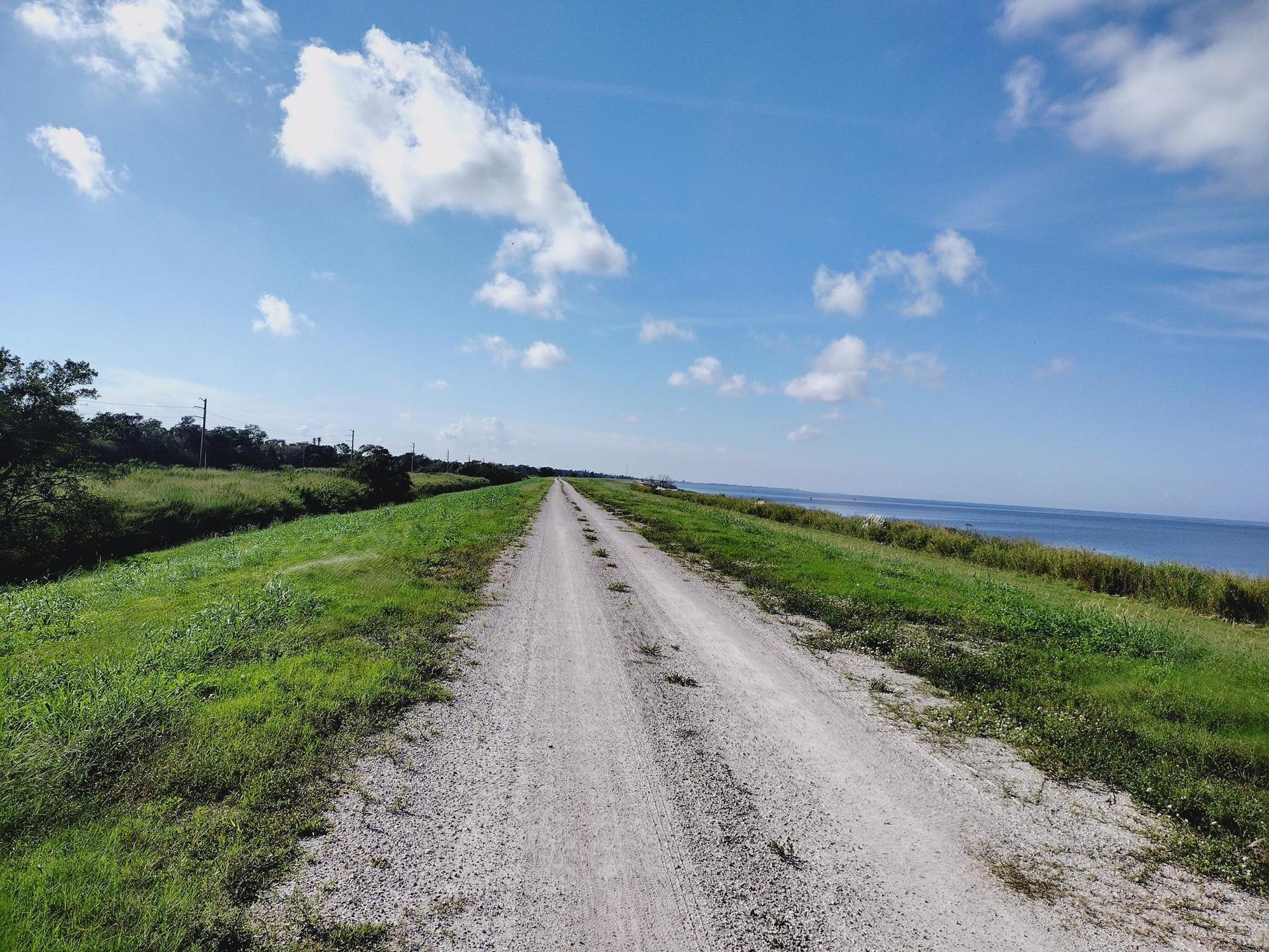 Long gravel road between green banks, leading towards a bright blue sky.
