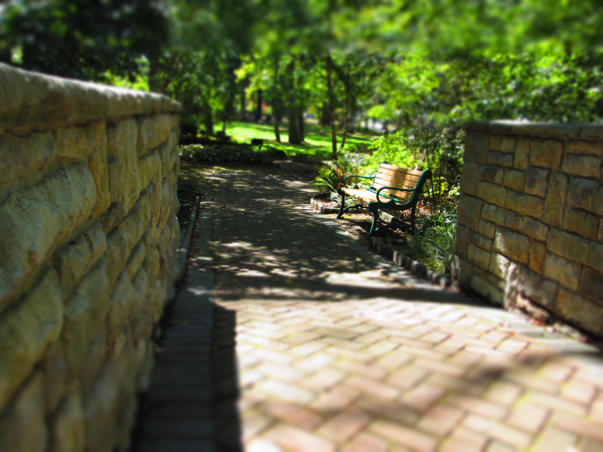 A brick path between stone walls leads to a park bench in dappled sunlight.