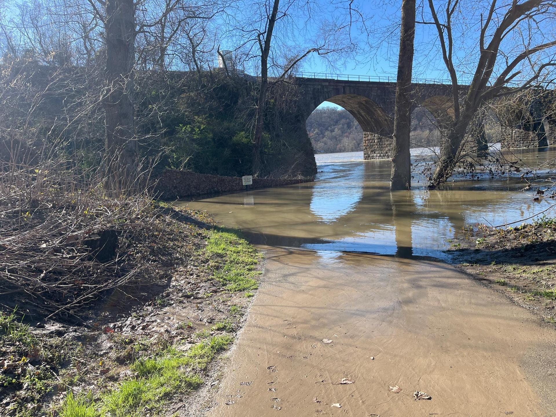Flooded dirt path by river, large stone arch bridge, bare trees.