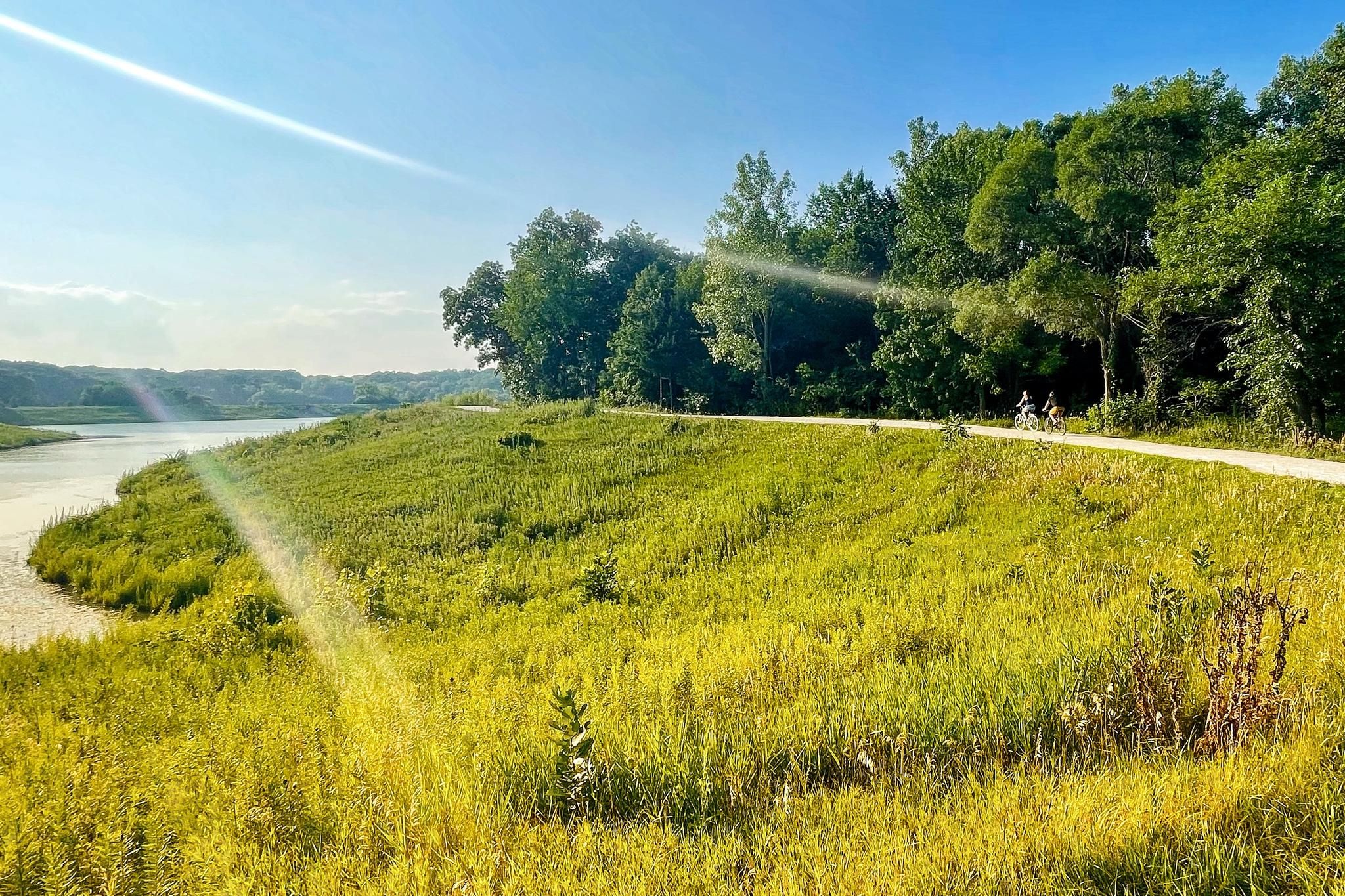 Sunny view of a river, grassy hill, and distant cyclists on a path. - Maple Lake and North Central DuPage Regional Trail Loop