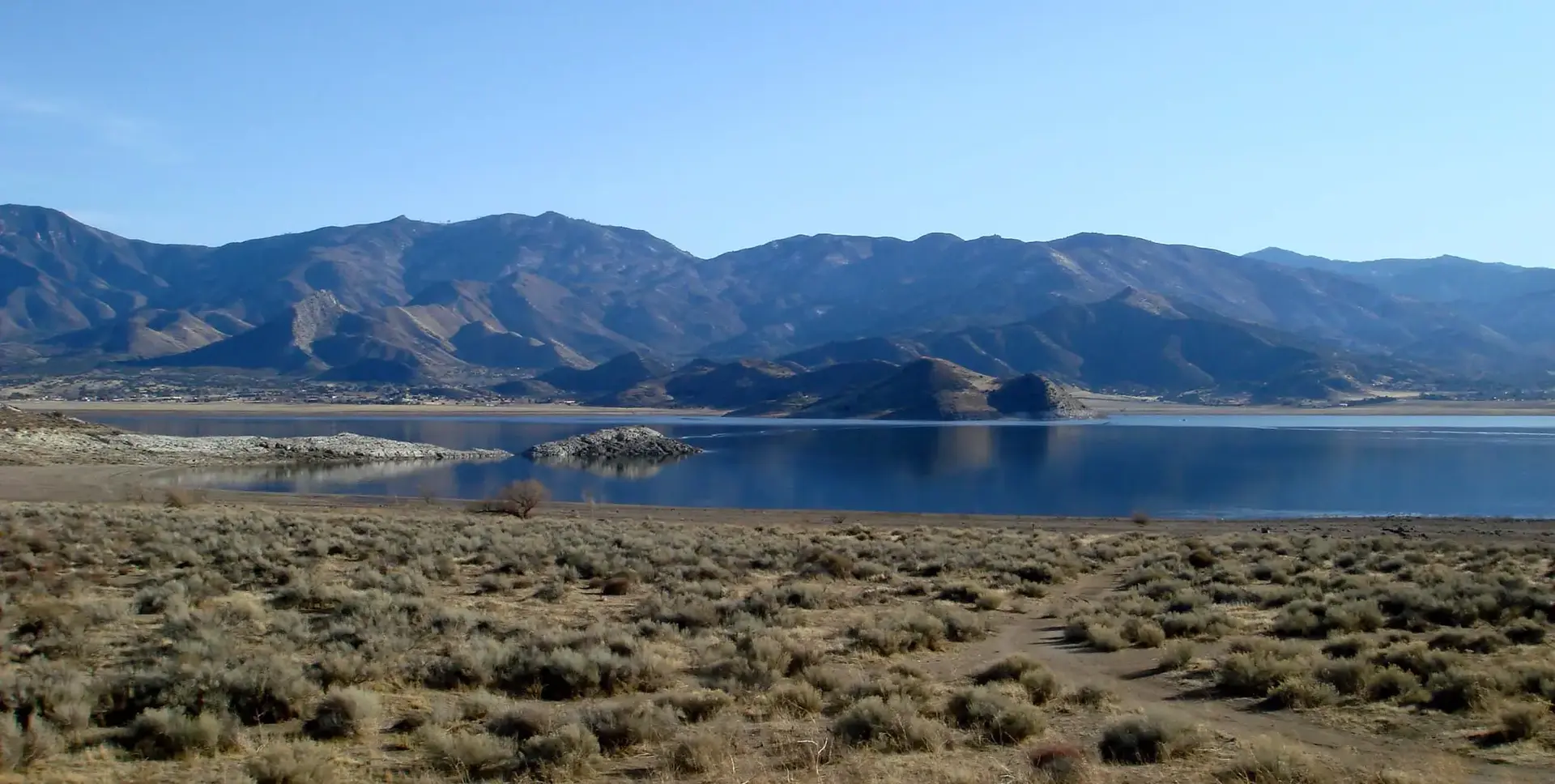 Calm lake with dry brush and rugged mountains under a clear blue sky.