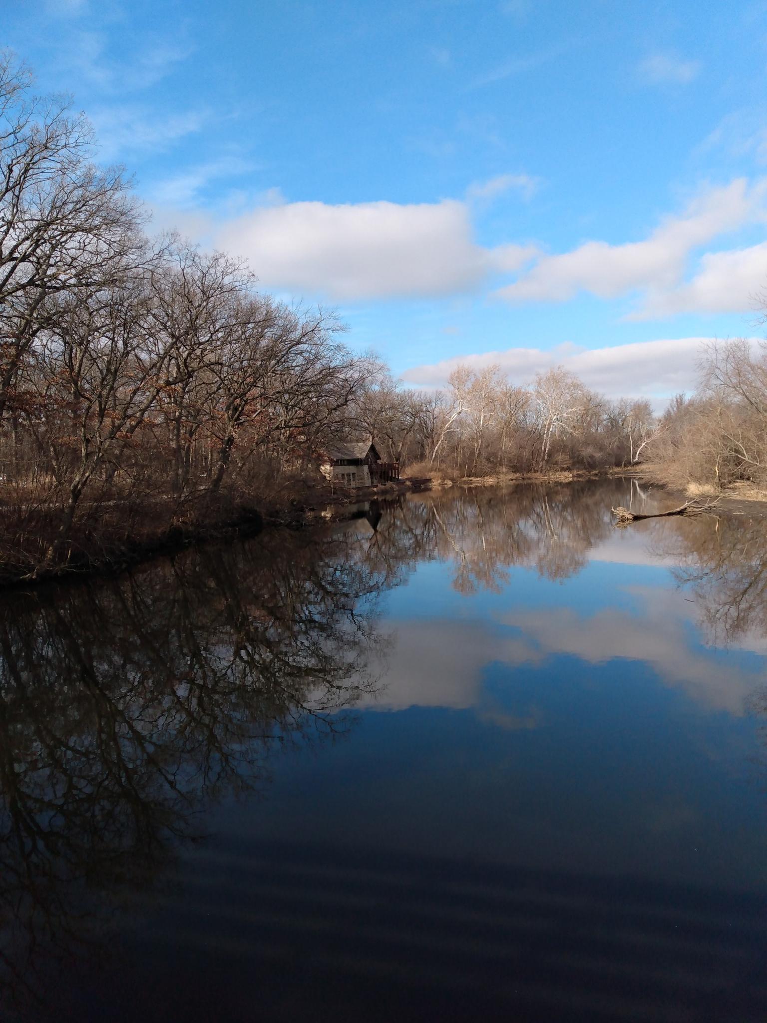 A calm river reflects bare trees and a blue sky with white clouds.