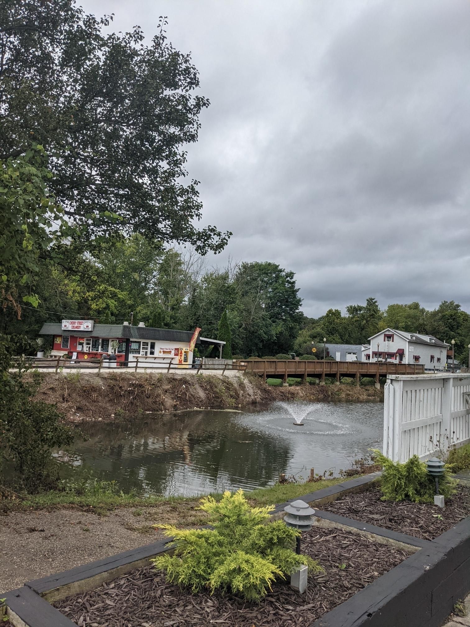 A pond with a fountain, buildings, and a wooden bridge under an overcast sky. - St Helena Heritage Park – Cherry Street Creamery loop from Canal Fulton