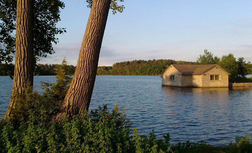 Stone house in a lake, with two large tree trunks in the foreground at sunset.
