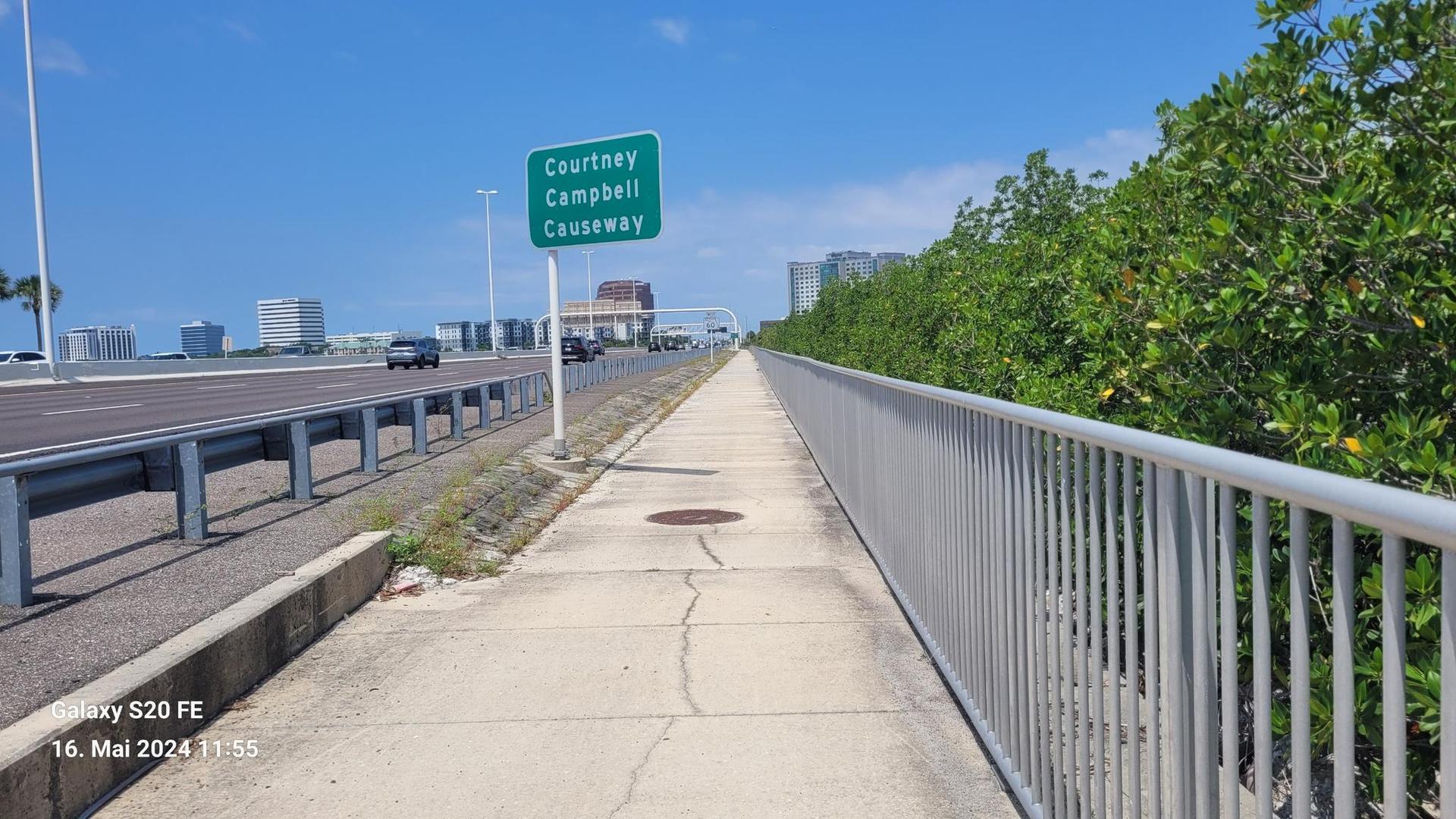 A sidewalk runs beside a highway with a green sign under a bright sky.