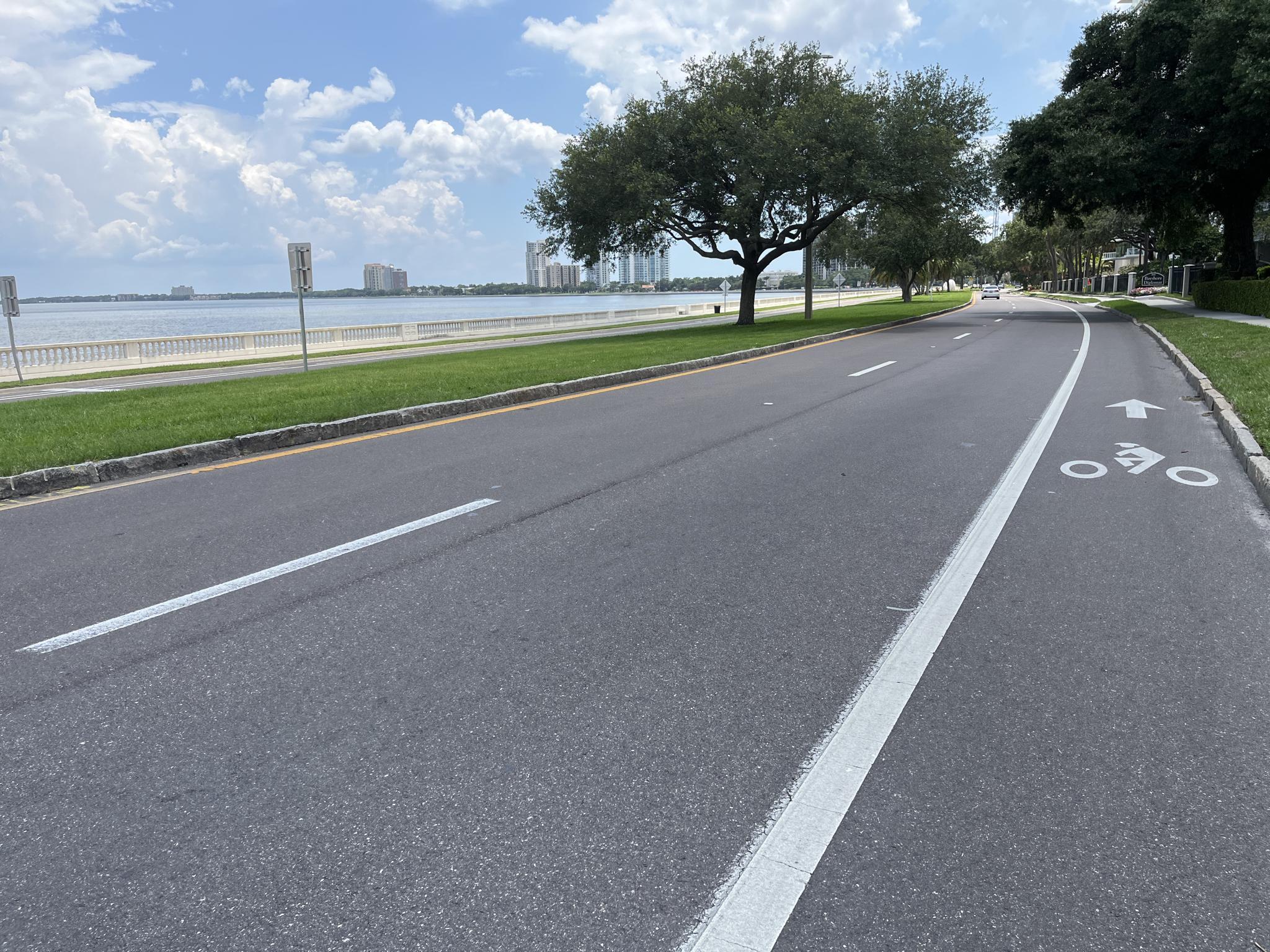 Coastal road with a bike lane, water, and distant buildings under a bright sky.